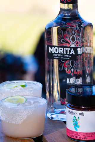 A bottle of Morita Chipotle Vodka stands on a wooden surface beside two margarita glasses with salted rims and lime wedges, plus a jar of fruit jam. The background is softly blurred outdoor scenery.