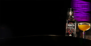 A bottle of Morita Karu Chipotle Vodka  stands on a dark surface next to a coupe glass filled with margarita with a chilli salt rim, set against a dark background with a purple light glow.