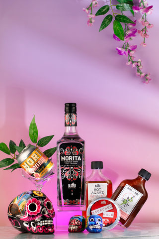 Bottles of Karu Morita Chipotle Vodka, Hot Agave, Hot Sauce and a can of Morita Grapegruit Soda on a pink background with hand-painted sugar skulls from Mexico.