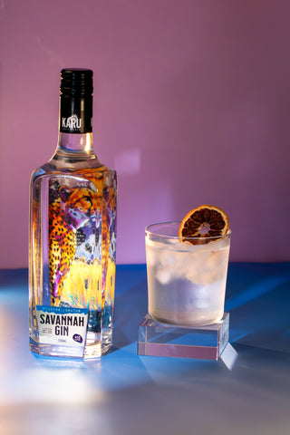A bottle of Karu Savannah Gin stands beside a rocks glass filled with a pale cocktail and a dried orange slice garnish, set against a purple-to-blue studio background.